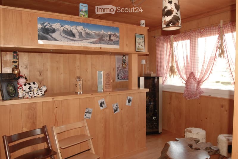 A cozy wooden-paneled room with a bar counter, chairs, a refrigerator, and various decorative items including cow figurines, books, and framed pictures.