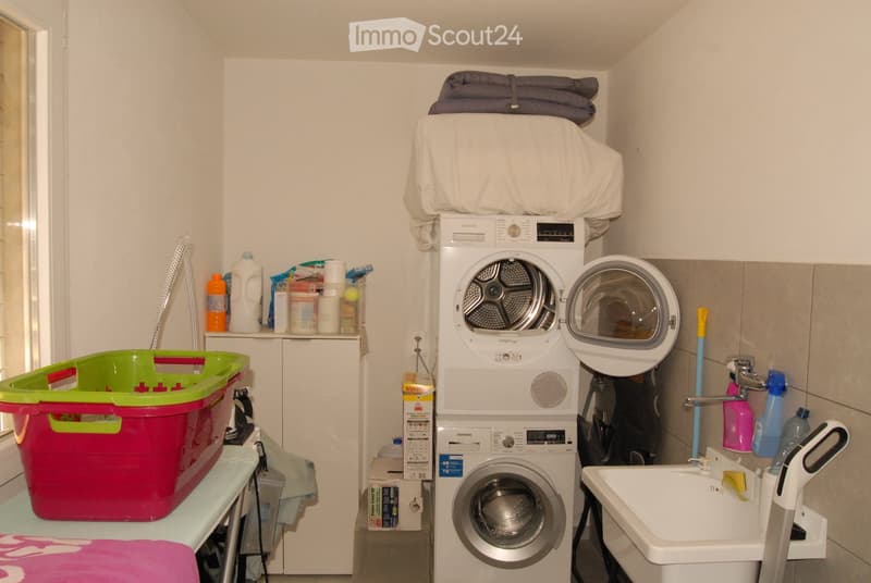 A compact bathroom with a stacked washing machine and dryer unit, a sink with a faucet, and various cleaning supplies and laundry baskets.