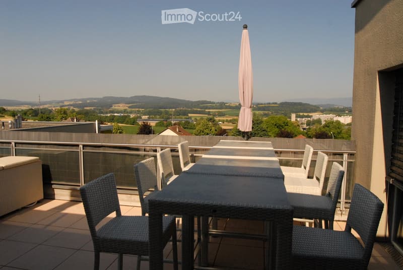 Balcony with outdoor furniture including a table, chairs, and a sofa, offering a scenic view of the countryside and cityscape.