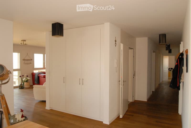 Bright hallway with wooden flooring, white walls, built-in white closet, and several doors leading to different rooms. Visible rooms include a living area with a red sofa, white armchair, and decorative items, as well as a kitchen area with hanging coats.