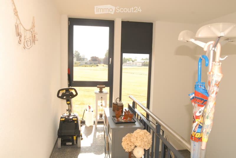Hallway with large windows, umbrella stand, flower vase, small table with decorative items, and an exercise bike.