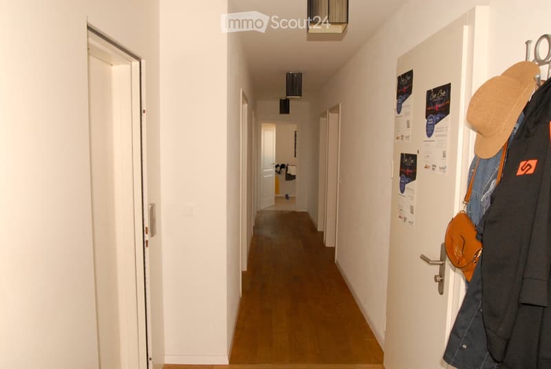 A narrow hallway with white walls, wooden flooring, and several doors leading to different rooms. Posters are displayed on the walls, and a coat rack with a hat and a bag is visible on the right side.