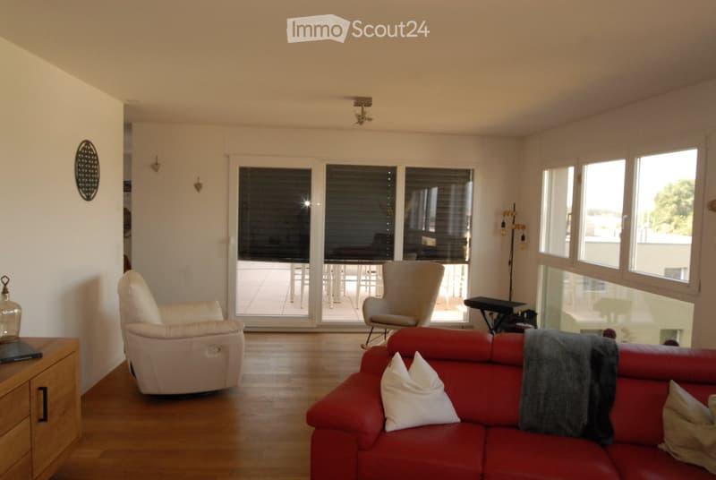 Bright living room with large windows and a sliding glass door leading to a balcony. Features a red sofa with white cushions, a beige armchair, a small table, and a floor lamp.