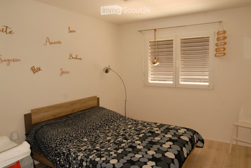 A modern bedroom with a wooden bed, patterned bedspread, floor lamp, and a white chair. The walls feature decorative words in gold lettering.