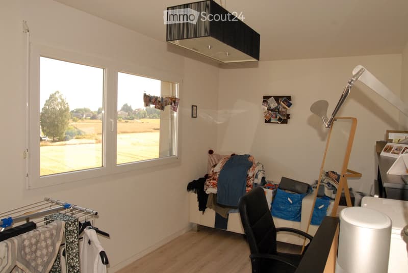 A small, furnished room with a bed, chair, lamp, and clothing rack. The room has large windows with a view of the street and a clothes drying rack near the window.