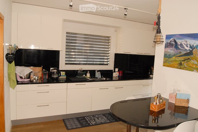 Modern kitchen with white cabinets, black countertops, a central island, and a round dining table with chairs. Features include a window with blinds, a painting on the wall, and various kitchen appliances and utensils.