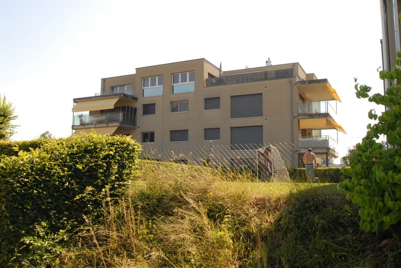 Multi-story residential building with balconies, large windows, and a raised ground floor. The building is surrounded by greenery and has a modern architectural style.