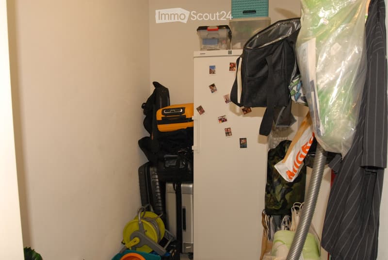 A small, organized storage closet with a refrigerator, suitcases, backpacks, a vacuum cleaner, storage bins, and a coat hanging on the wall.