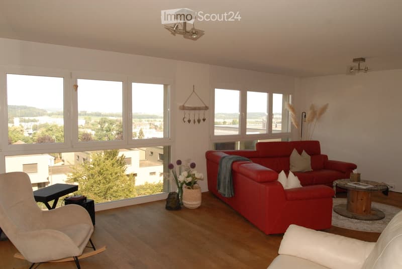 Modern living room with large windows offering a scenic view, featuring a red sofa, a white armchair, a coffee table, and decorative items including a vase with flowers and a hanging ornament.