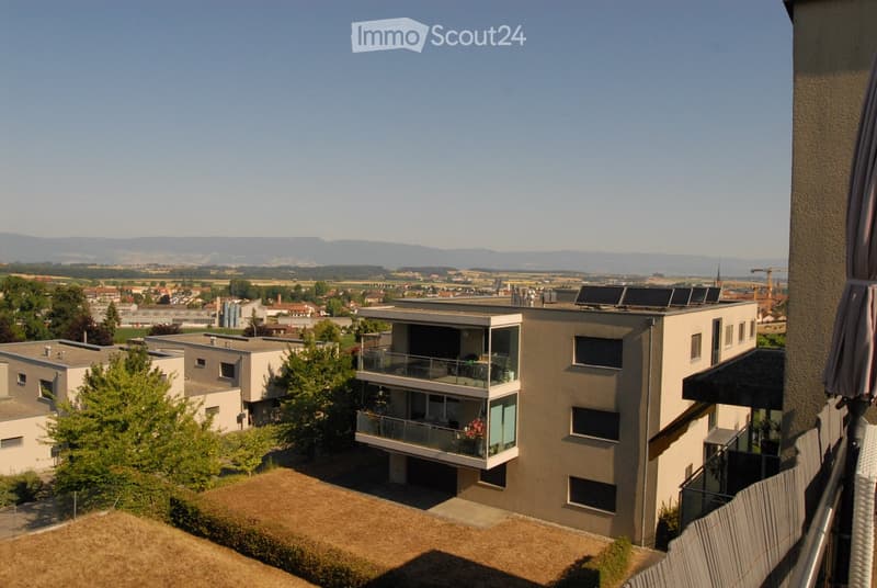 Modern multi-story residential building with balconies, solar panels on the roof, and a scenic view of a town and distant mountains.