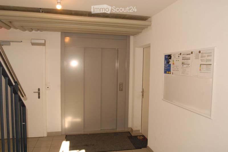 A clean, modern hallway with a staircase on the left, an elevator door in the center, and a bulletin board on the right wall.