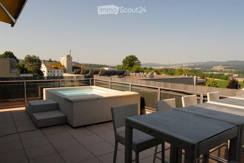 A modern rooftop terrace with a hot tub, a table, and several wicker chairs, overlooking a scenic countryside view.