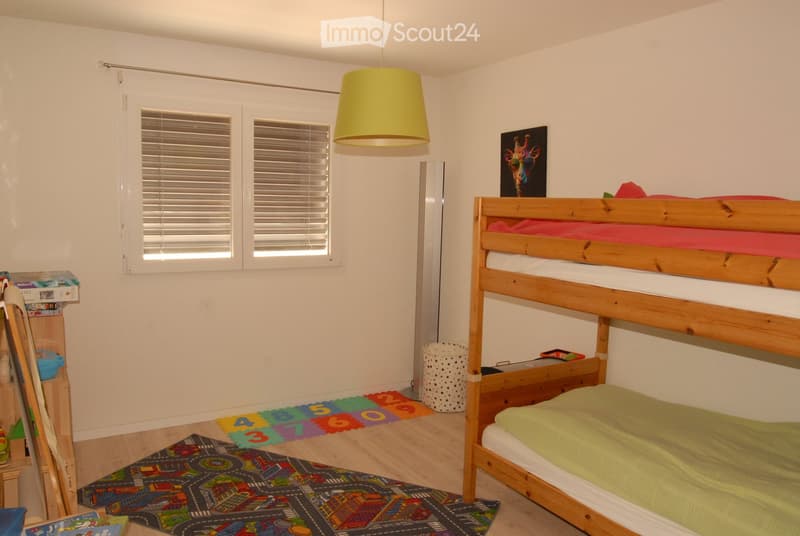 Bright bedroom with a wooden bunk bed, colorful play mat, and a hanging lamp. The room features white walls, wooden flooring, and a window with blinds.