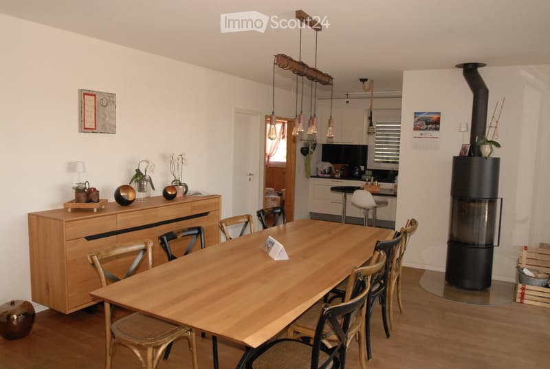 Bright, open-plan living and dining area with wooden flooring, a large wooden dining table surrounded by chairs, a wooden cabinet with decorative items, a modern black wood-burning stove, and a visible kitchen area with pendant lights and a small island.