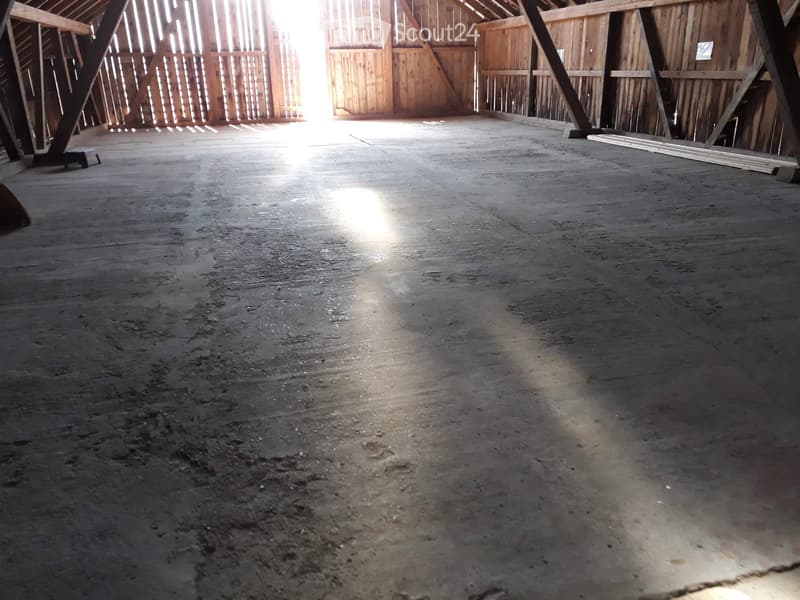 Empty barn interior with wooden beams, concrete floor, and natural light from windows.