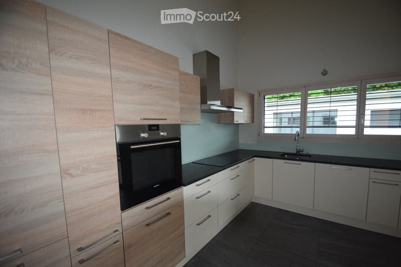 Modern kitchen with light wood cabinets, stainless steel oven, black countertop, and a window with blinds.