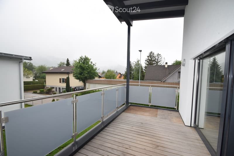 Modern balcony with wooden decking, glass railing, and metal balustrade, overlooking a residential area with houses and trees.