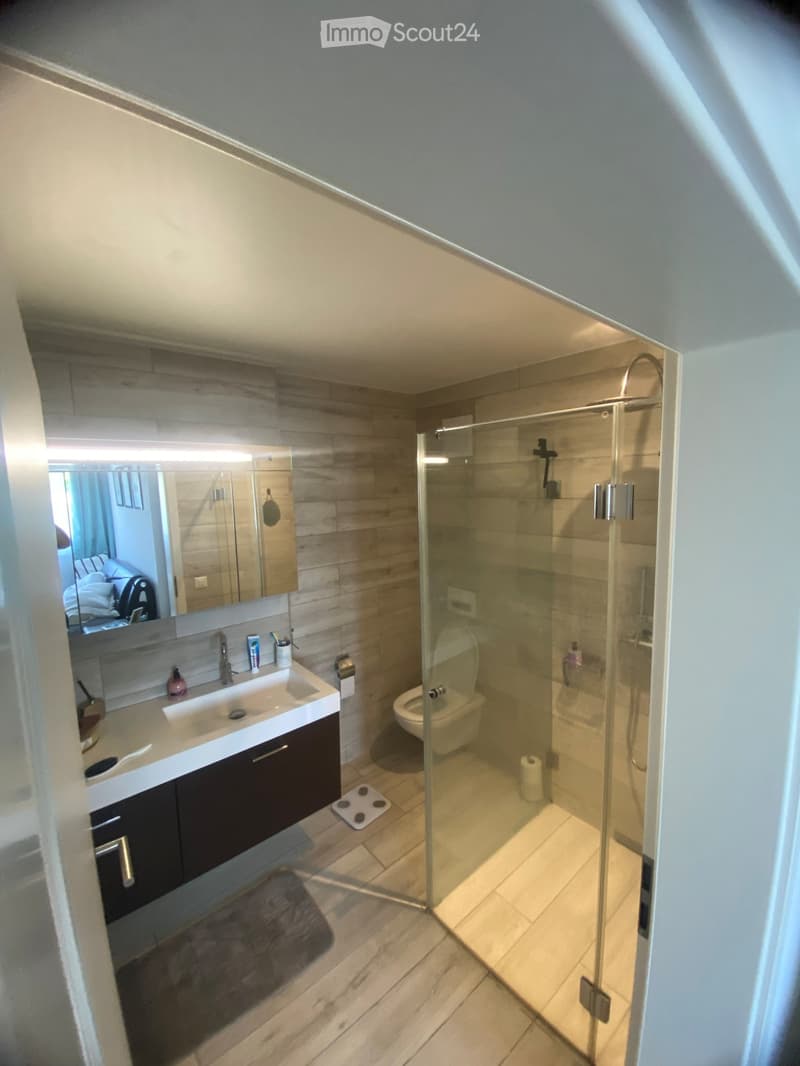 Modern bathroom with a glass-enclosed shower, a white sink with a dark cabinet, a toilet, and a large mirror with built-in lighting. The floor is tiled, and the walls feature a wood-like finish.