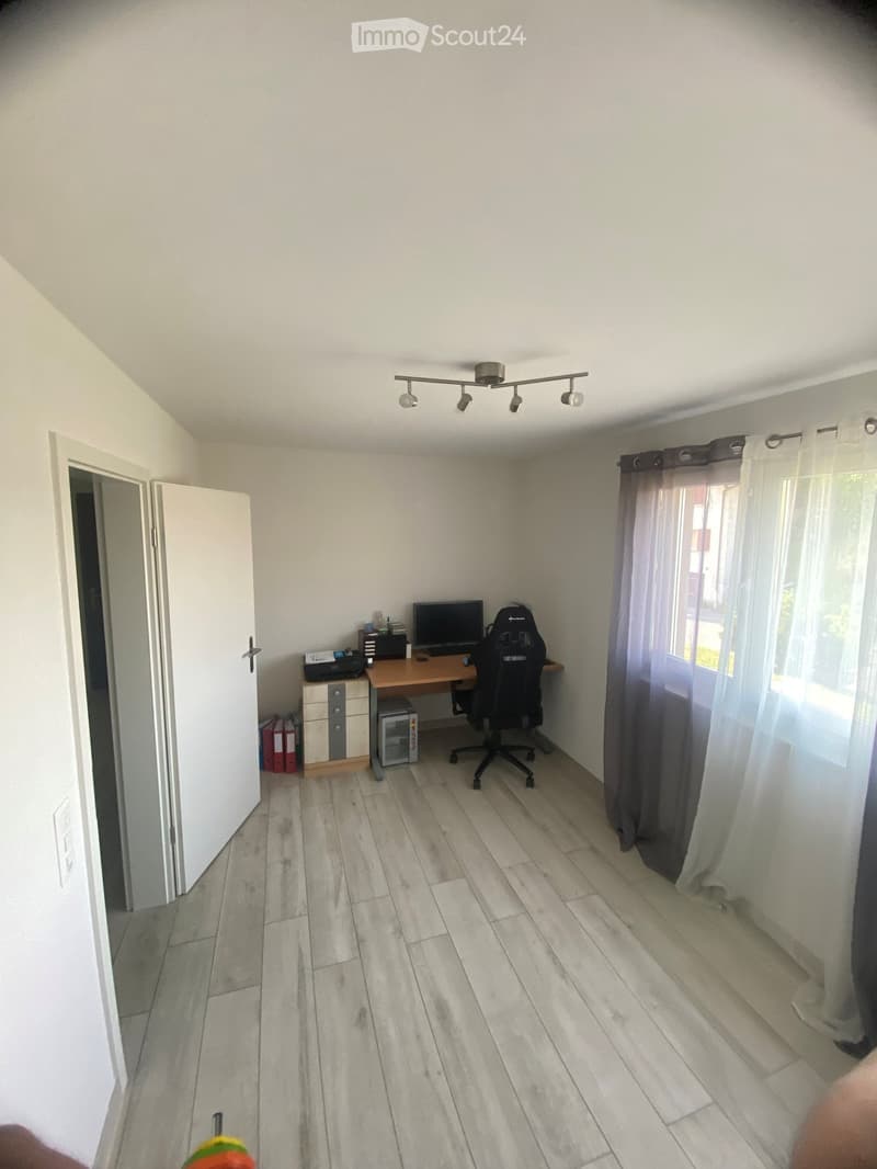 A furnished room with a wooden desk, computer, office chair, printer, and cabinet. The room has light-colored wooden flooring and white walls.
