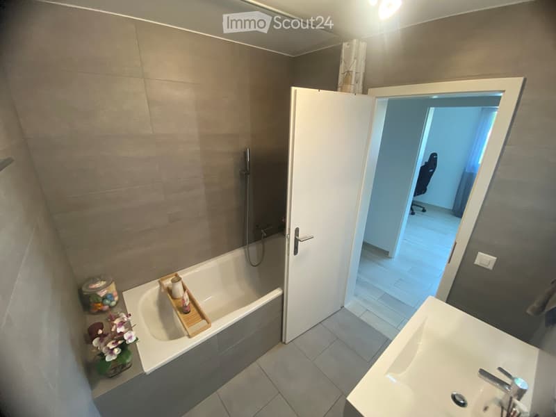 Modern bathroom with a bathtub, sink, and a wooden tray holding toiletries. A flower arrangement is placed next to the bathtub, and the room features tiled flooring and walls.