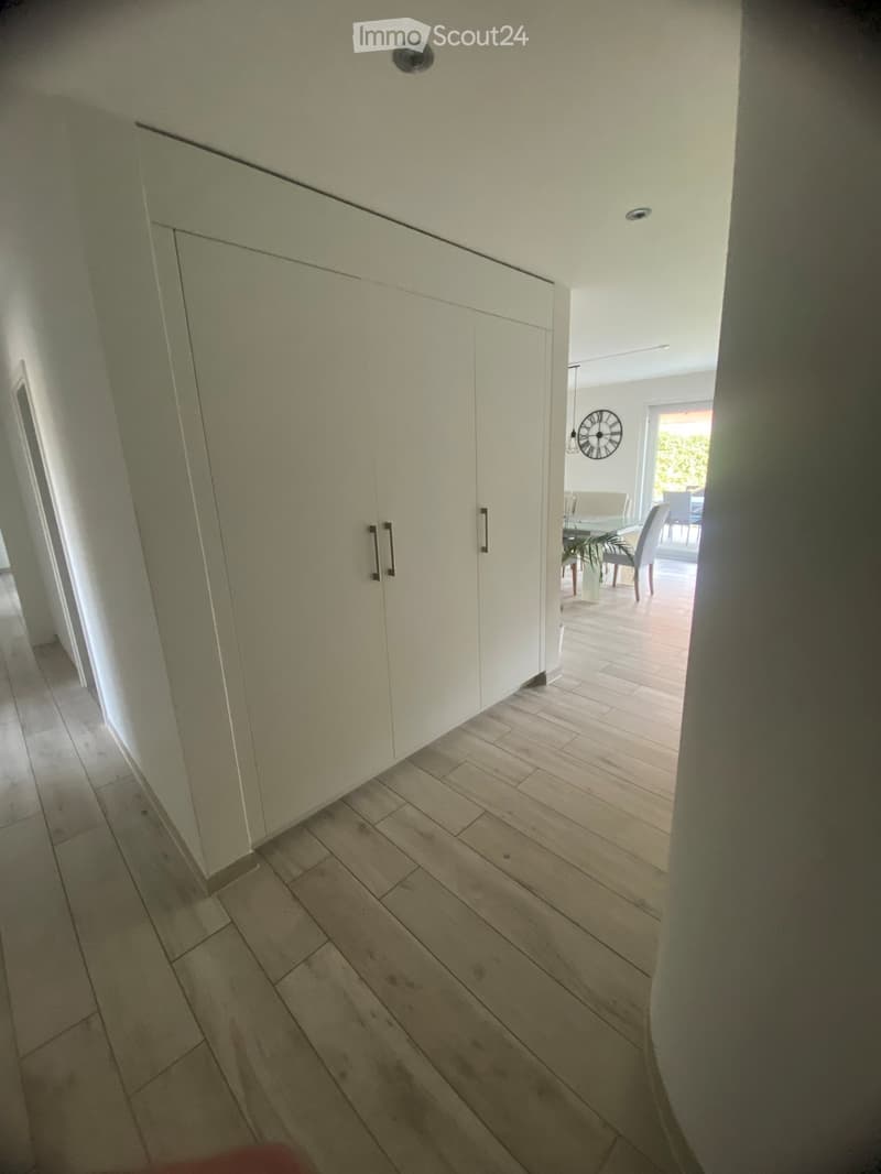 A well-lit hallway with light-colored wooden flooring and white walls, featuring built-in storage cabinets with sleek handles and a visible open area leading to a dining space with chairs and a table.