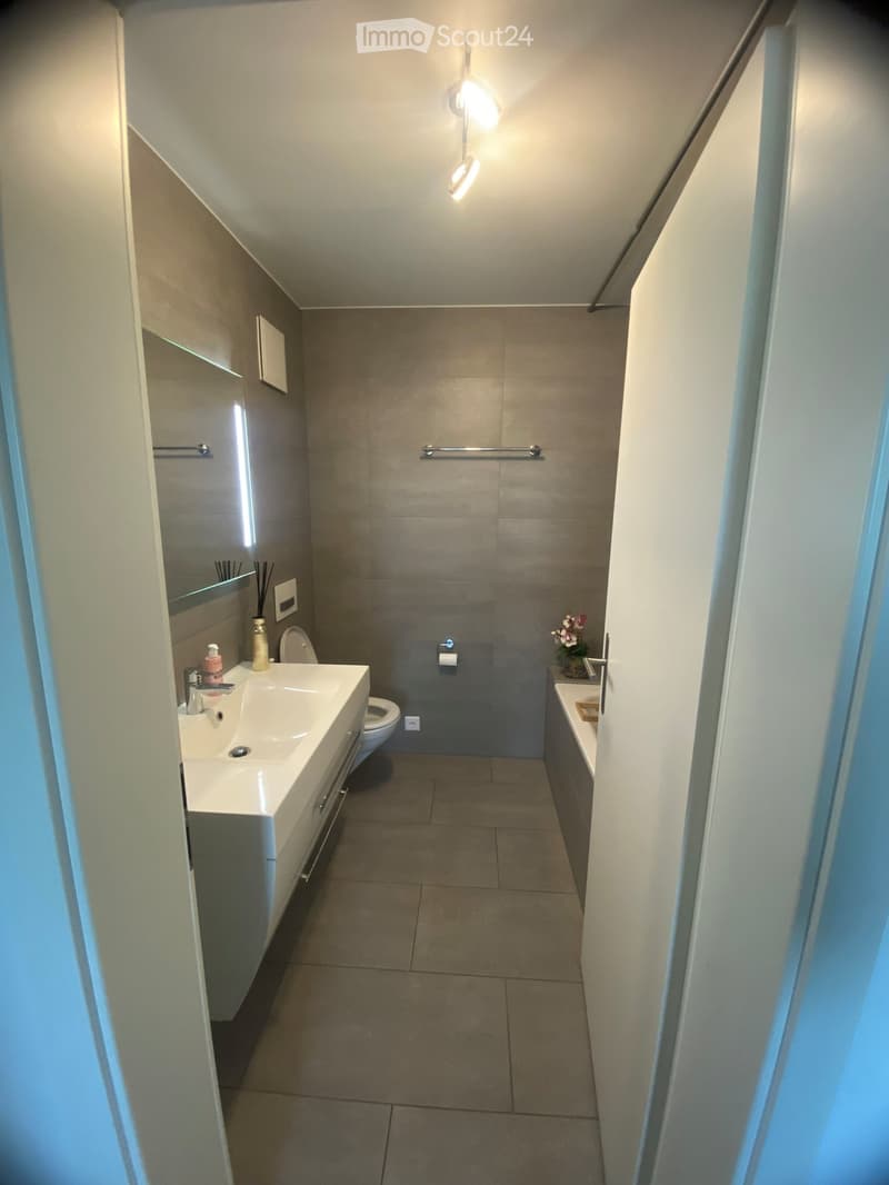 Modern bathroom with a double sink, toilet, mirror, and a small bathtub. Features include a towel rack, a flower vase, and a sleek, contemporary design with gray tiles and white fixtures.