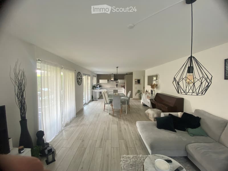 Open-plan living and dining area with light wooden flooring, grey sofa, coffee table, dining table with chairs, piano, and decorative items.