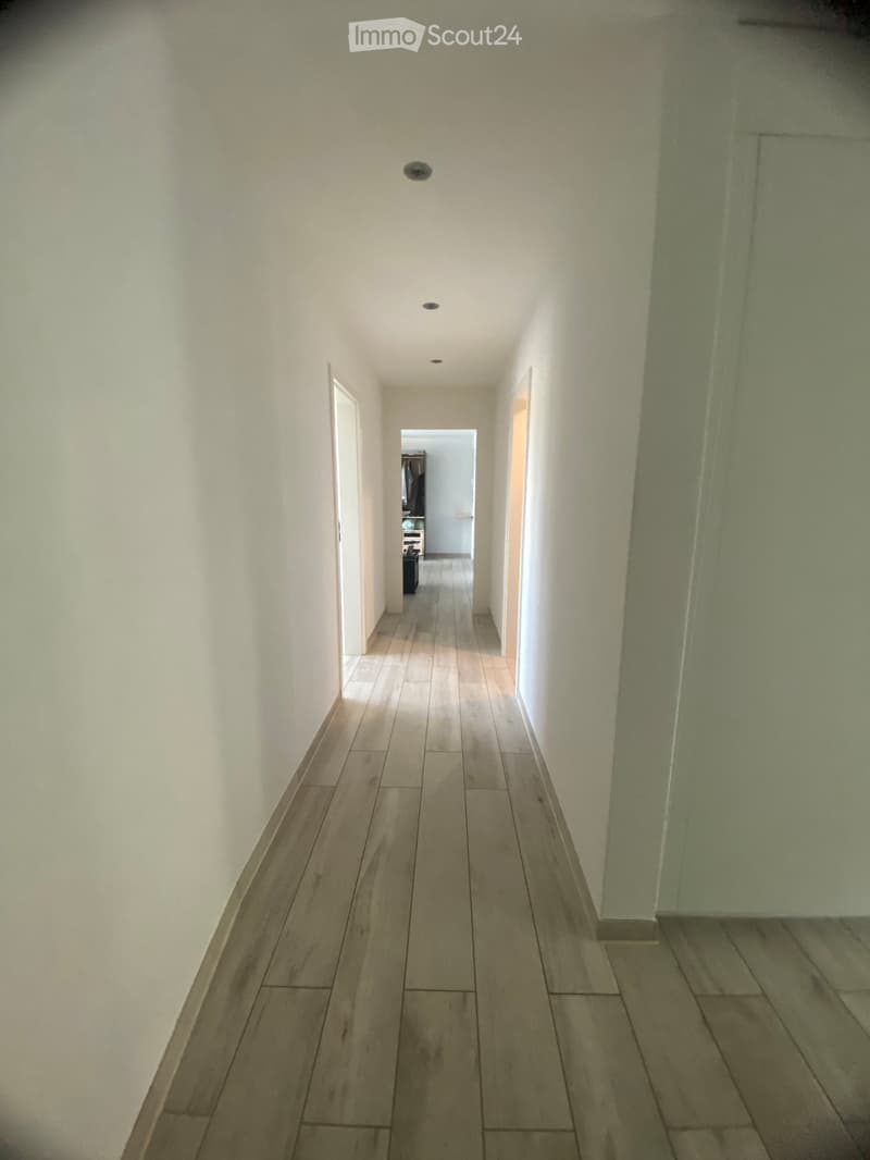 A long, narrow hallway with light-colored wooden flooring and white walls, featuring recessed ceiling lights and doors leading to adjacent rooms.