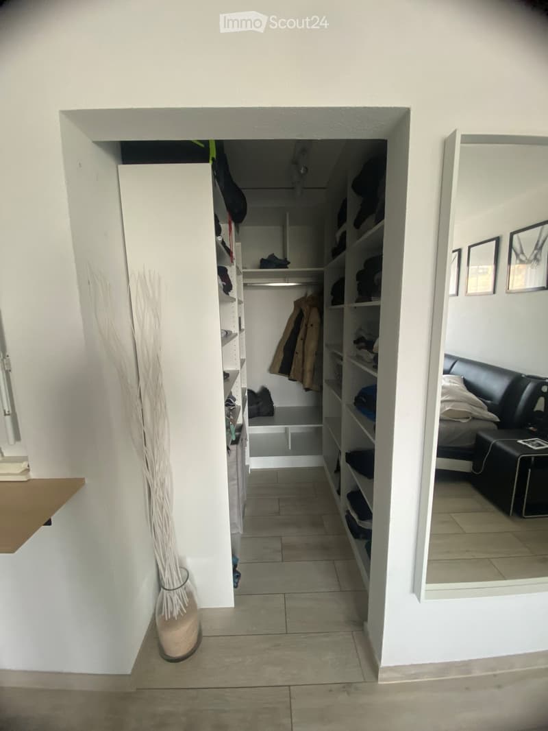 A narrow hallway with tiled flooring, leading to a closet with shelves and a bedroom area with a bed and nightstand.