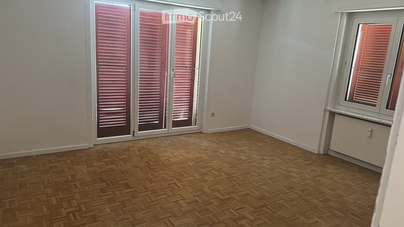 Empty room with wooden flooring, white walls, large window with shutters, and a sliding glass door leading outside.