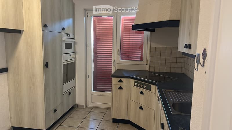 Modern kitchen with light wooden cabinets, stainless steel appliances, black countertops, and a window with blinds.