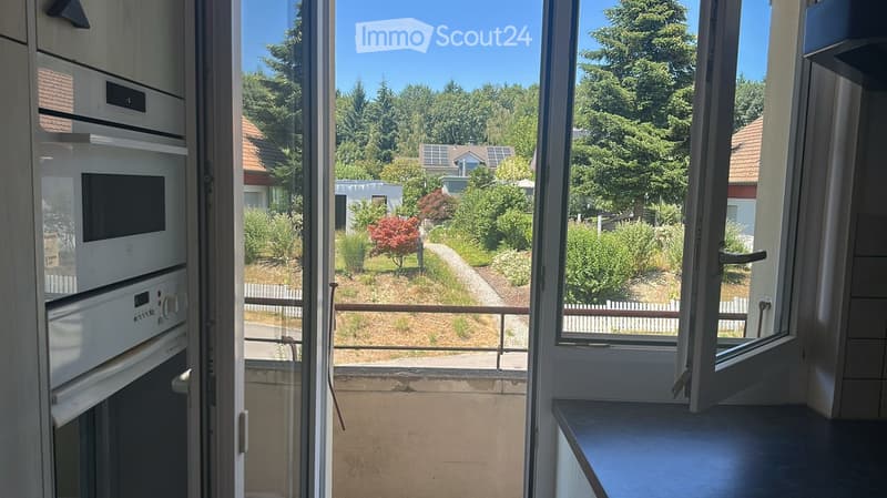 Balcony with open sliding doors, overlooking a garden with trees and houses in the background.