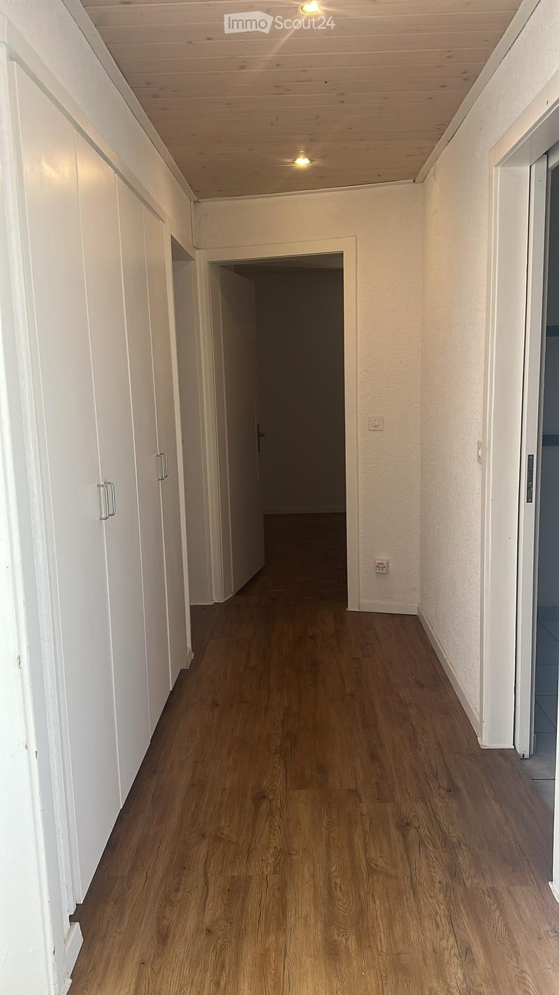 A narrow hallway with wooden flooring, white walls, and white cabinetry on one side. The hallway leads to an open doorway at the end.