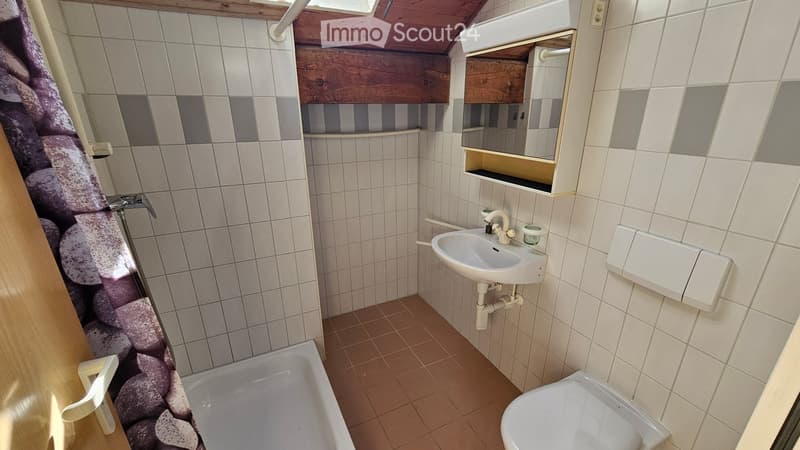 Bathroom with white tiled walls, a sink, toilet, bathtub, and a mirror cabinet. The floor is tiled in a warm brown color, and there is a wooden shelf above the sink.