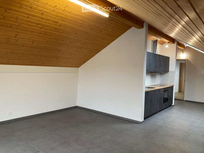 Empty room with wooden slanted ceiling, grey tiled floor, and a small fitted kitchen area with cabinets and appliances.