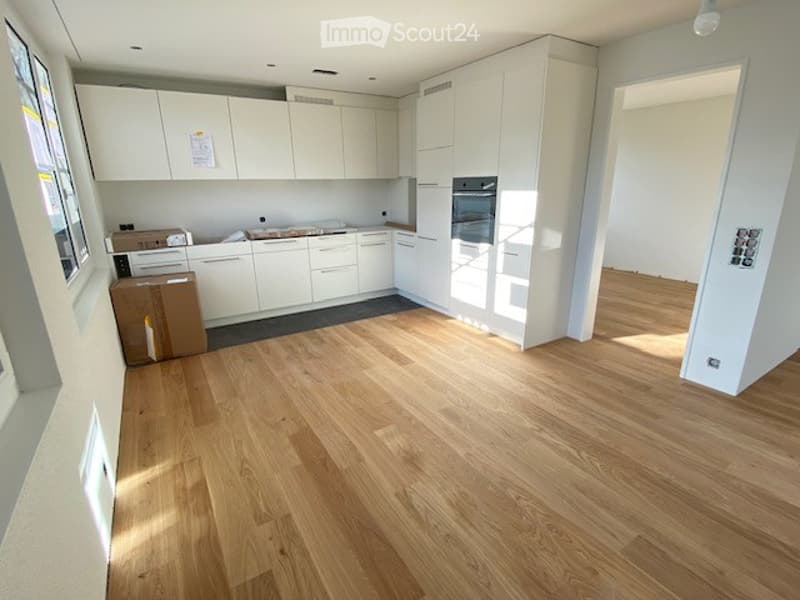 Empty kitchen with white cabinets, wooden flooring, and natural light from windows. No furniture or appliances are present.