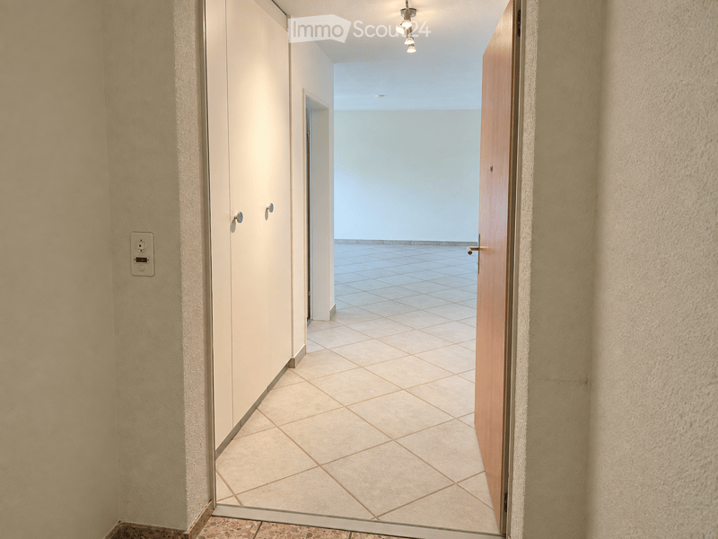 Empty hallway with tiled floor, light-colored walls, and recessed ceiling lights. Features a closed wooden door on the right and a partially open door leading to another room on the left.