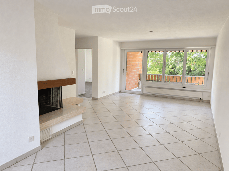 Empty room with tiled floor, white walls, fireplace with wooden mantel, large windows with wooden frames, and a sliding glass door leading to a balcony.