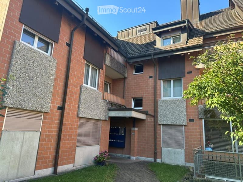 Multi-story brick apartment building with multiple windows, balconies, and a small garden area in front.