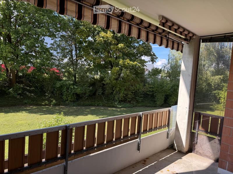 Balcony with wooden railing, striped awning, and view of a green lawn and trees.