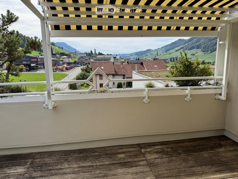 Balcony with wooden decking, white railing, and retractable awning, overlooking a town with houses and green hills.