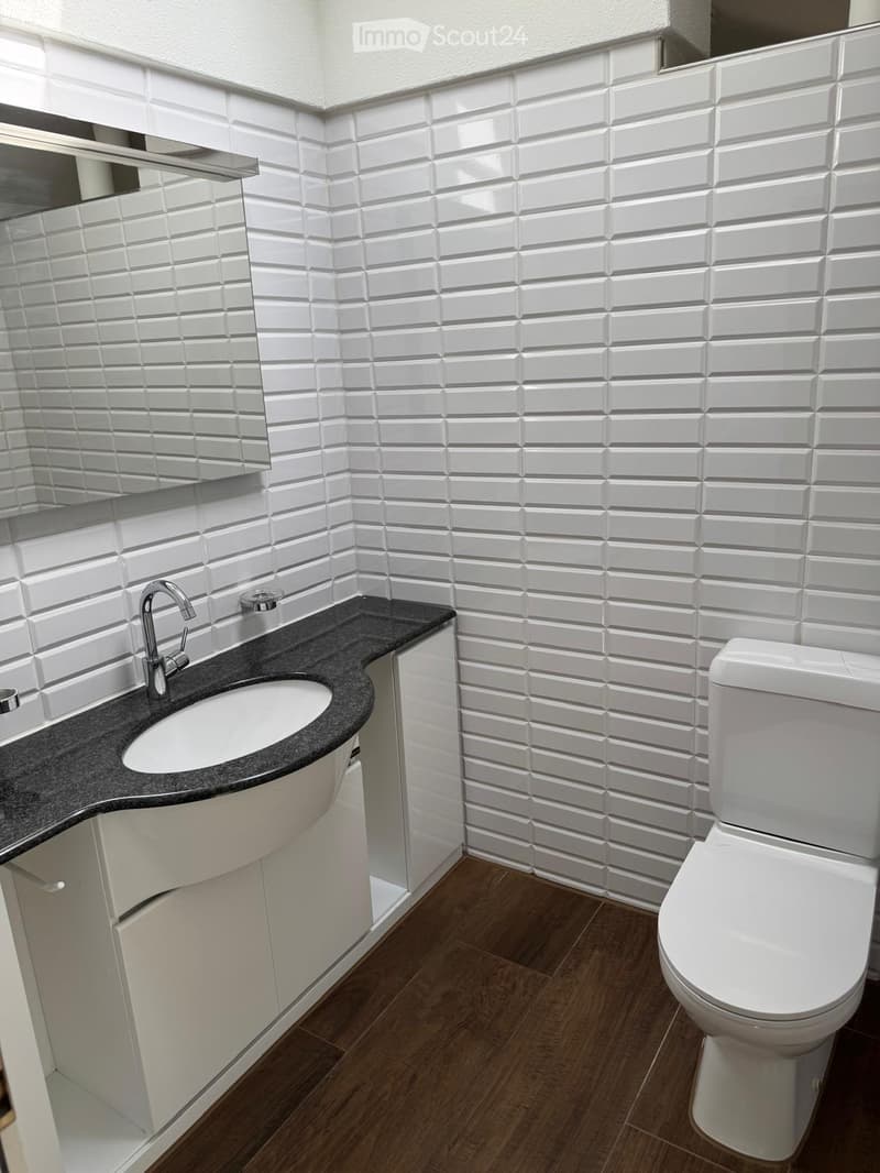 Modern bathroom with white tiled walls, dark countertop, white sink, chrome faucet, and white toilet.