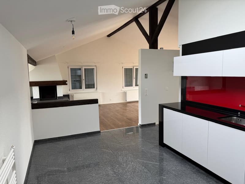 Open-plan living and kitchen area with exposed wooden beams, modern white cabinets, dark countertops, and large windows. The space is unfurnished, showing only the structural elements and built-in features.