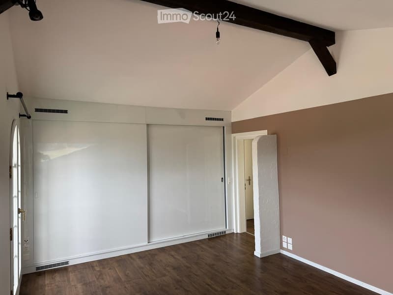 Empty room with wooden flooring, white walls, and exposed wooden beams on the ceiling. Features include a large white sliding door, a small doorway leading to another space, and a textured white column.