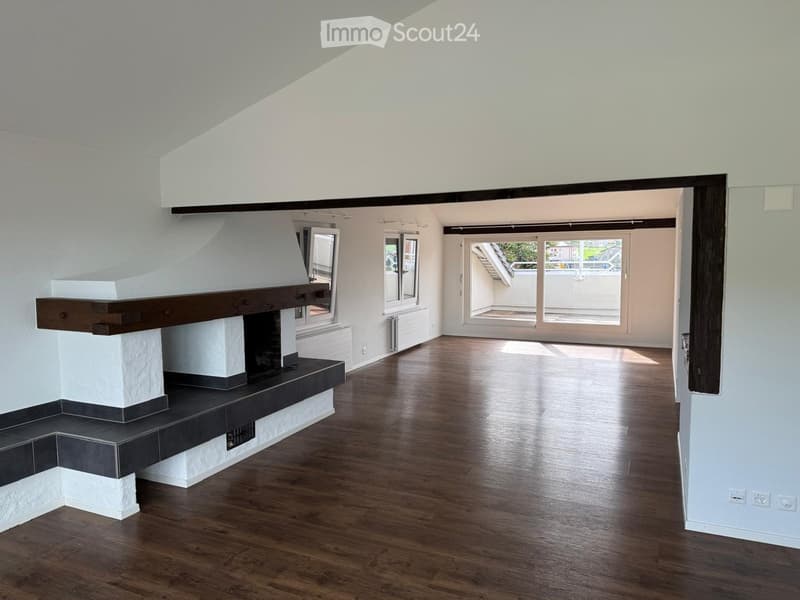 Empty room with wooden flooring, white walls, and a fireplace. Large windows and a sliding glass door lead to a balcony or terrace area.