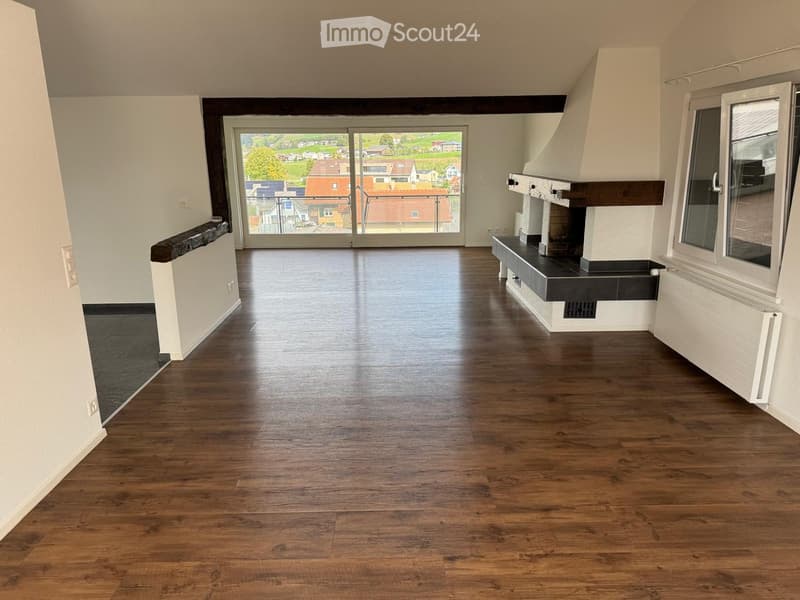 Empty room with wooden flooring, white walls, and a fireplace. Large windows and sliding doors lead to a balcony with a view of the neighborhood.