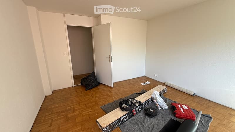Empty room with wooden flooring, white walls, and a partially open door leading to another space. Some construction materials and tools are scattered on the floor.