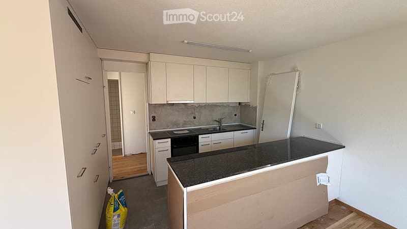 Modern kitchen with white cabinets, black countertop, sink, and oven. Wooden flooring and a partially visible doorway leading to another room.
