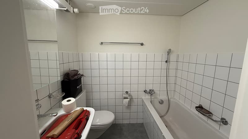 A small bathroom with white tiled walls, a bathtub, a toilet, a sink with a mirror, and a towel rack. The space includes a showerhead and a towel on the floor.