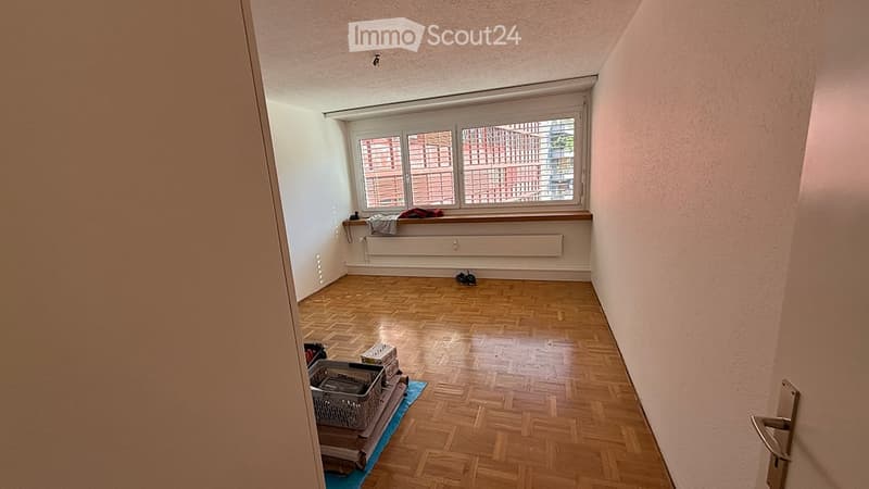 Empty room with wooden flooring, large window, radiator, and a few small items near the window and door.